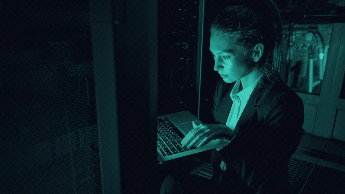 A technician, seen in profile, at her laptop in front of a server rack in low light.